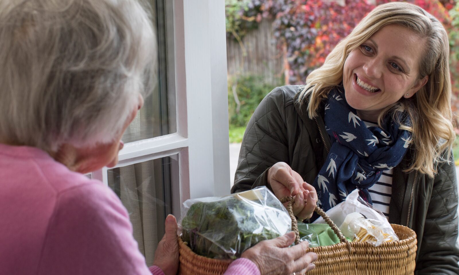 A carer smiling warmly with an older woman, holding a basket of fresh shopping at the front door.